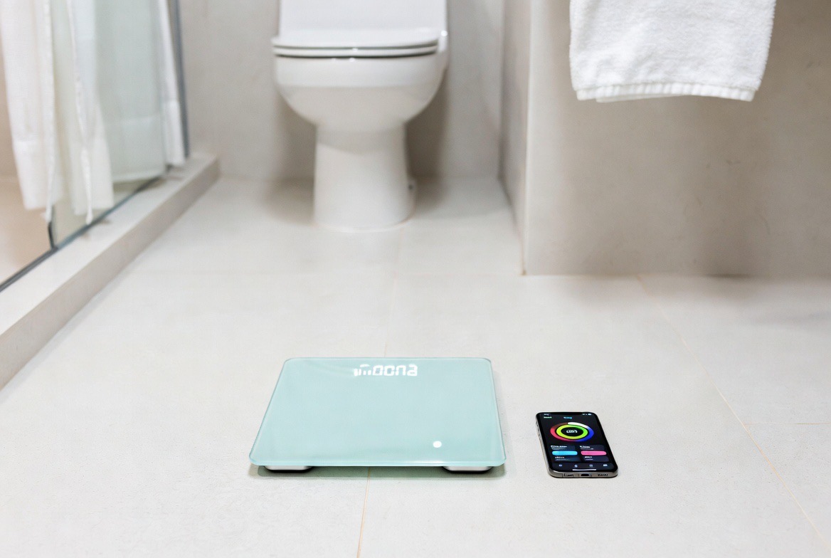 A modern smart bathroom scale beside a phone showing a body-composition dashboard — the scale is the clear visual anchor