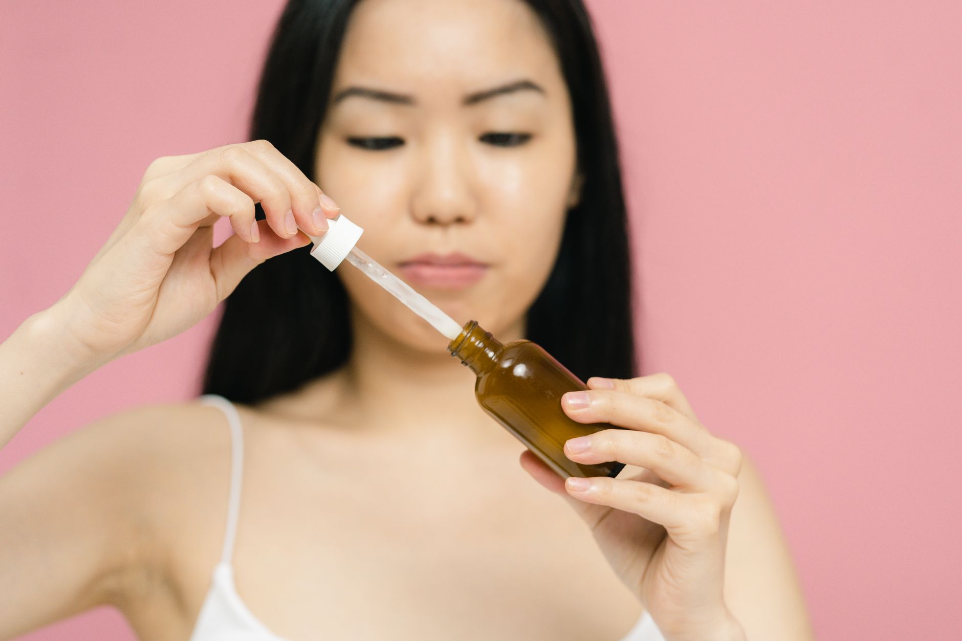 A woman holding an amber glass skincare serum bottle and drawing a clear essence into a dropper, against a soft pink background &mdash; the K-beauty serum aesthetic snail mucin is sold in