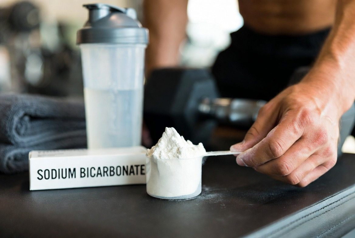 An athlete's hand holding a scoop of white sodium bicarbonate powder beside a box labeled SODIUM BICARBONATE, a shaker bottle, dumbbell and gym towel in a bright training setting