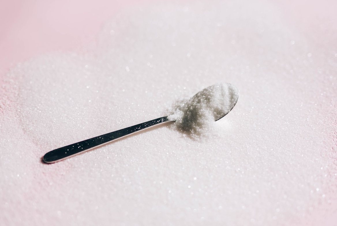 A metal spoon holding a heap of white crystalline granulated sweetener resting on a bed of the same sugar-like crystals, illustrating erythritol and xylitol sugar alcohols
