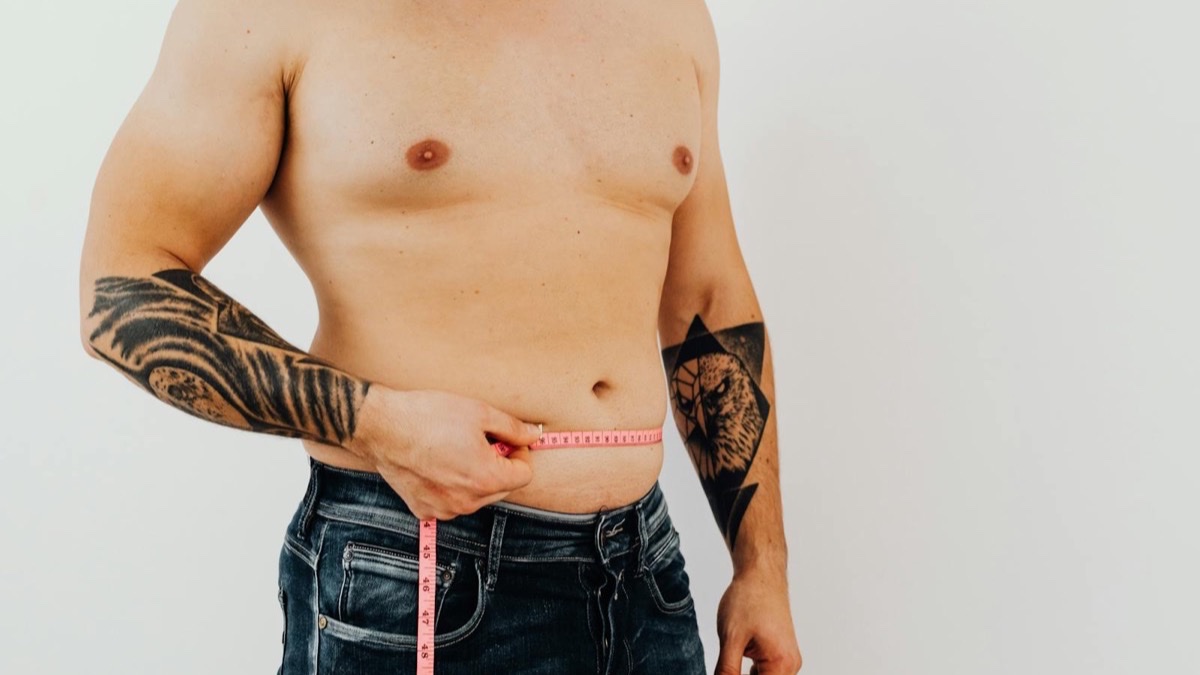 Shirtless man measuring his waist with a tape measure — assessing midsection and visceral belly fat, the target of tesamorelin