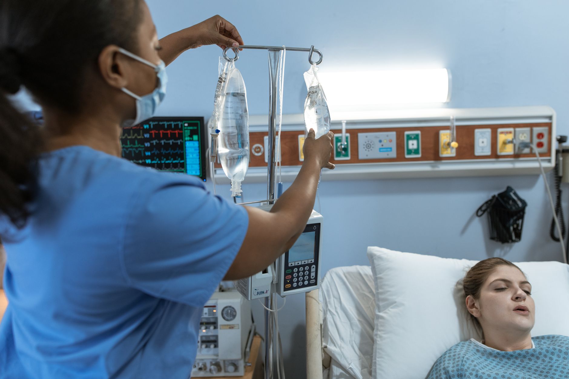A nurse in a mask hangs an intravenous fluid bag on a drip stand beside a hospitalized patient resting in a clinical bed, with a cardiac monitor in the background
