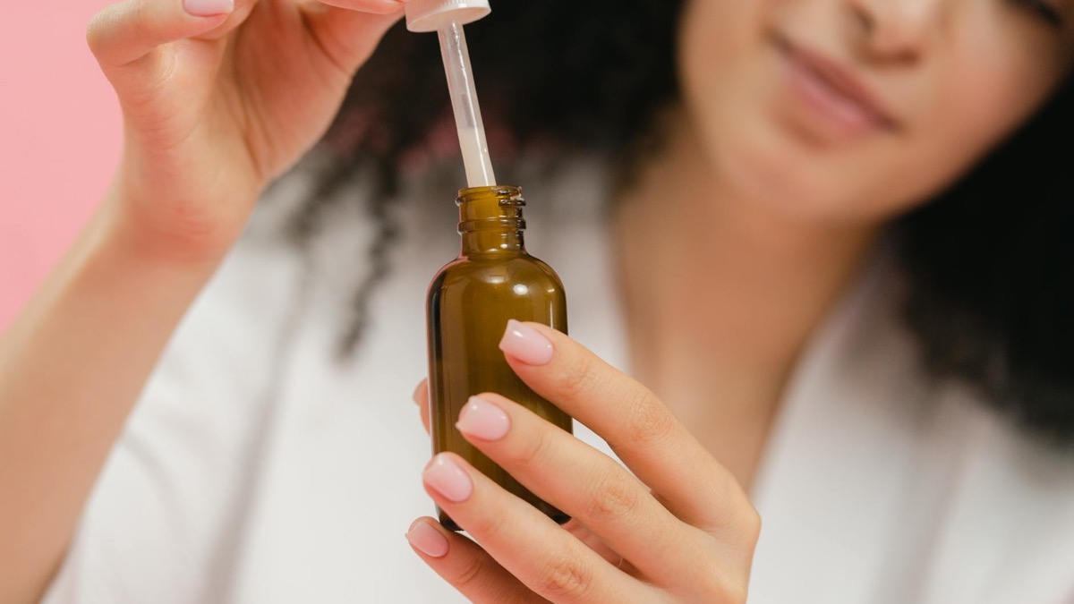 Woman lifting the dropper from an amber glass serum bottle against a soft pink background — applying a topical skincare treatment
