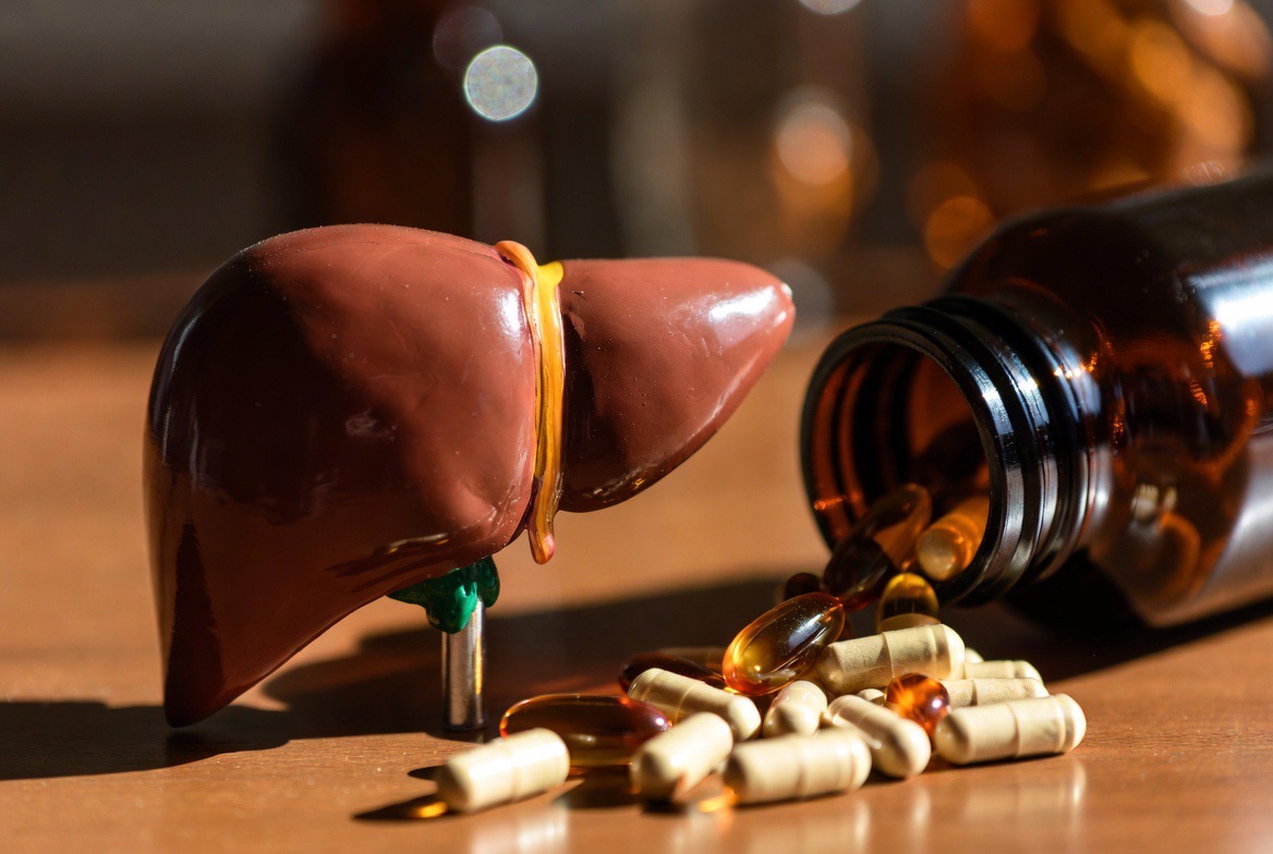 Amber-coloured bile-acid supplement capsules spilled from a dark glass bottle beside an anatomical liver model on a neutral editorial surface