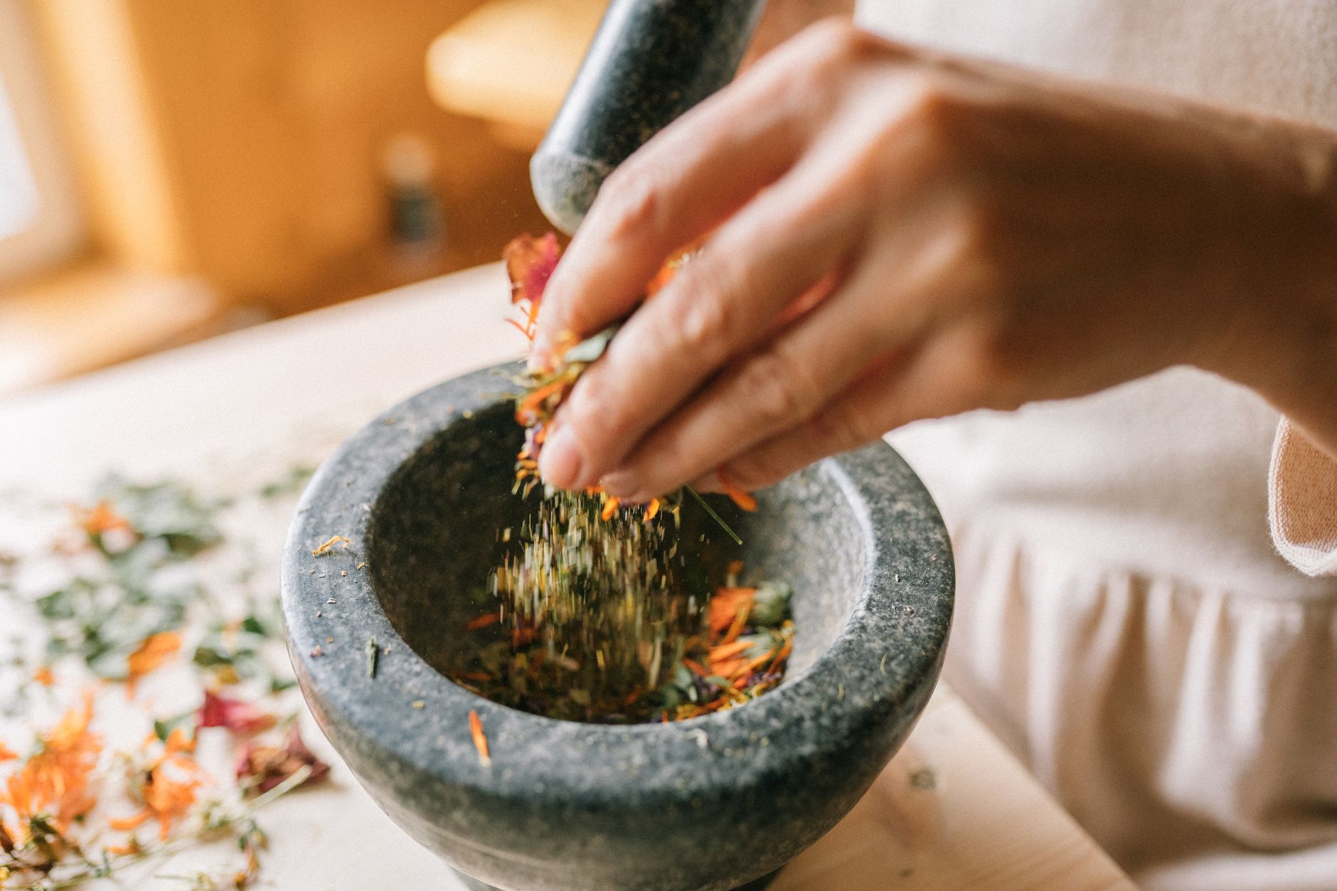 Hands grinding dried botanical herbs and flowers with a stone mortar and pestle on a wooden table in warm natural light, evoking traditional herbal sleep-aid preparation