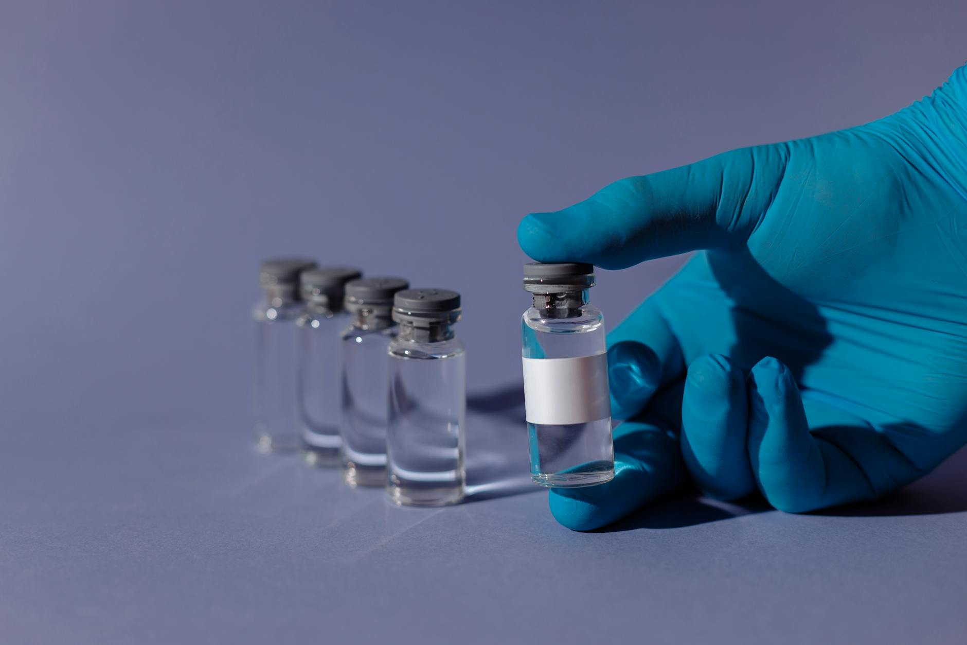 Row of clear glass injection vials with rubber stoppers handled by a gloved hand — peptide dosing and protocol context