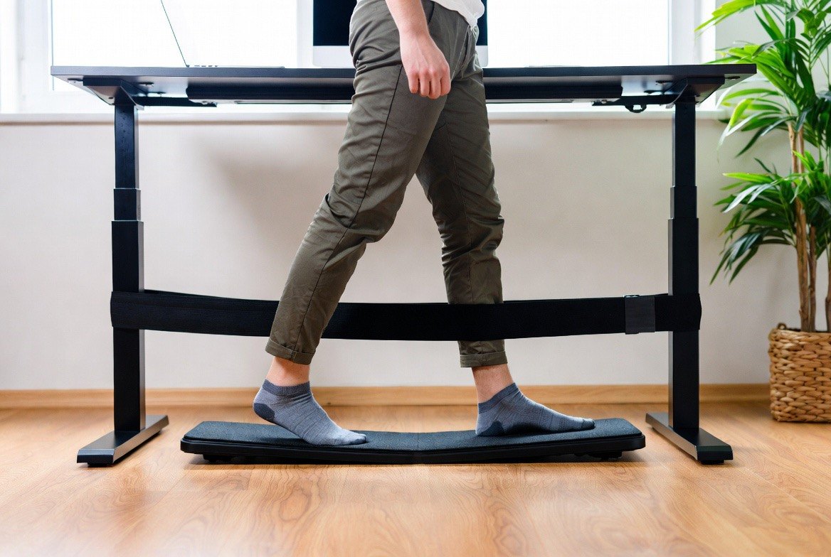 A person in socks walking on a slim under-desk walking pad beneath a black standing desk with a laptop on top, a potted plant and window behind — the low-intensity desk walking a walking pad adds to a workday