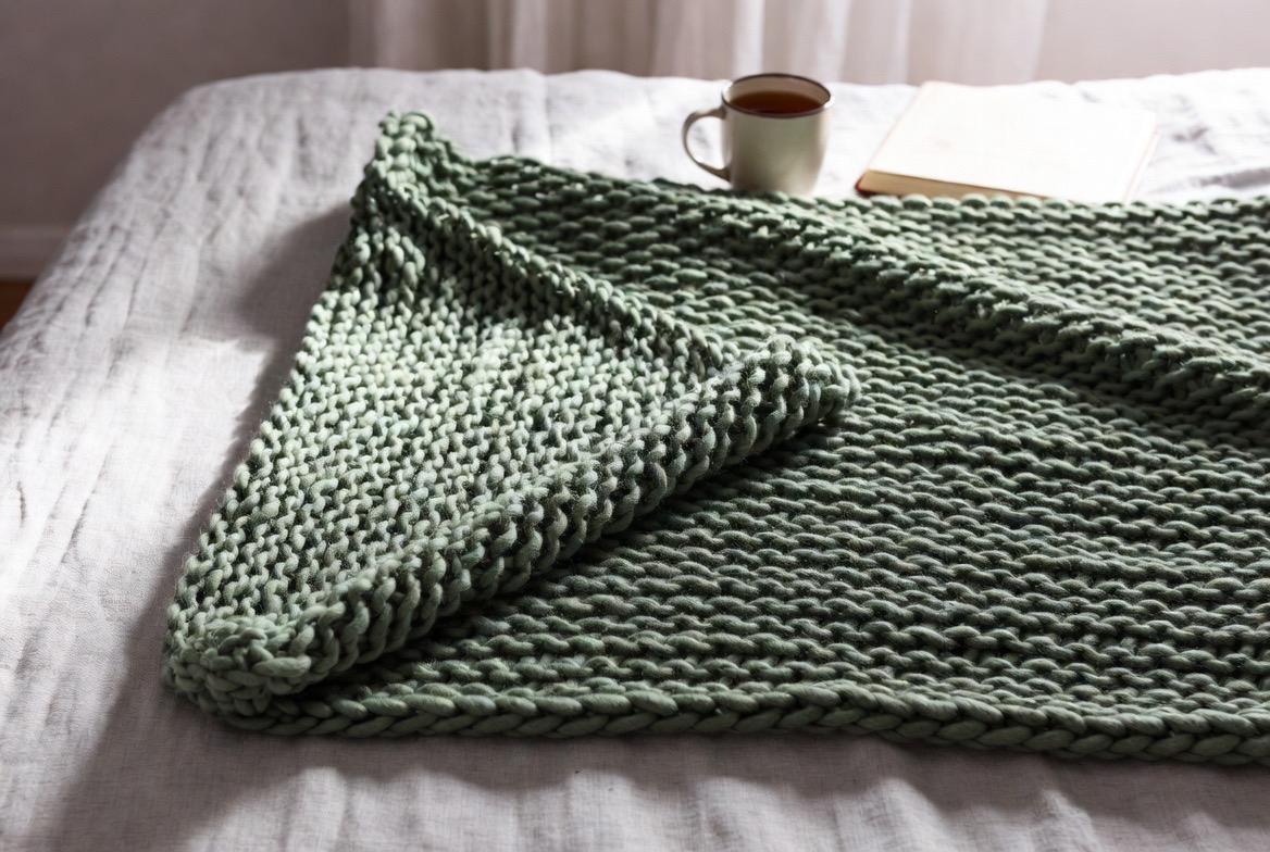 A chunky knit weighted blanket draped over a neatly made bed in soft morning light, the heavy textured blanket the clear focus of the frame