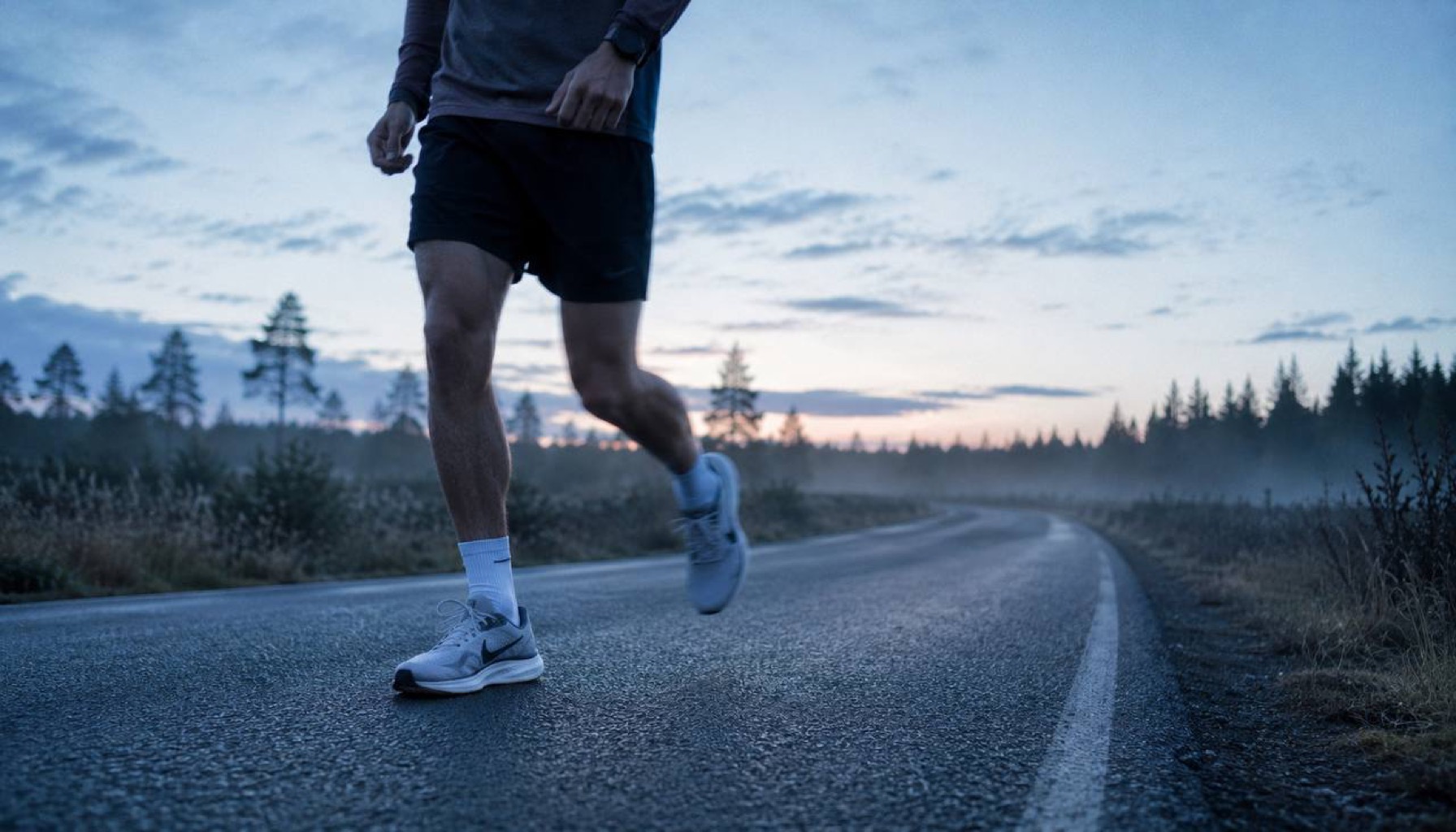 Runner's legs at steady aerobic pace on empty asphalt road in cool blue-grey dawn light with light mist